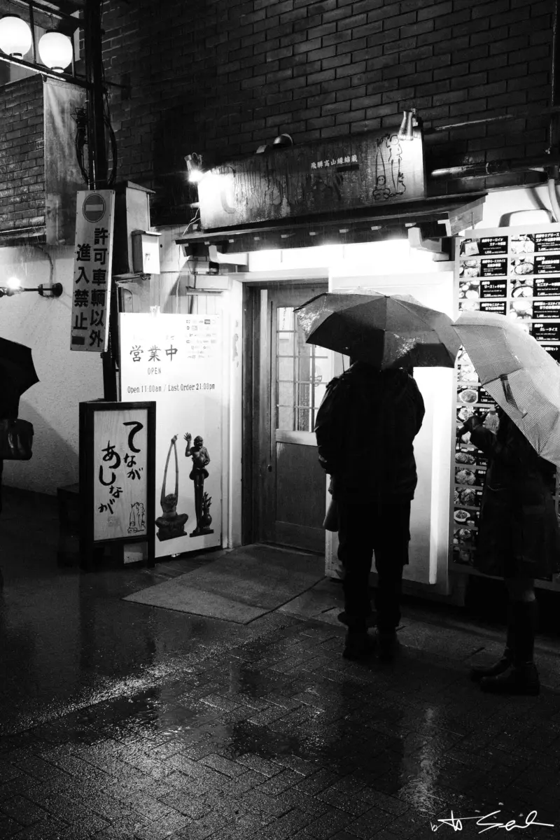 Menschen mit Regenschirmen vor einem Lokaleingang bei Nacht, Schwarz-Weiß